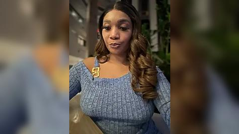 Media: Video of a confident Black woman with wavy brown hair, wearing a blue knit sweater, and a yellow handbag. She stands in a blurred indoor setting.