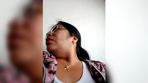 Media: Video of an Asian woman with medium skin tone, long black hair, wearing glasses, a white tank top, and a pink camo jacket, looking contemplative against a plain white background.