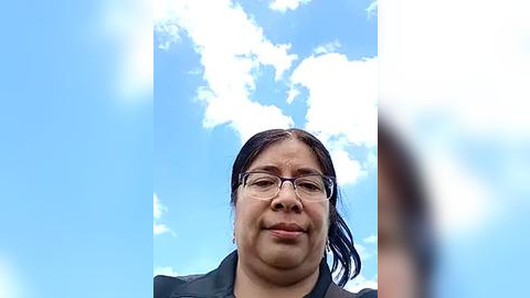 Media: A video of a middle-aged woman with medium skin tone, wearing glasses and a black jacket, standing against a bright blue sky with fluffy white clouds.