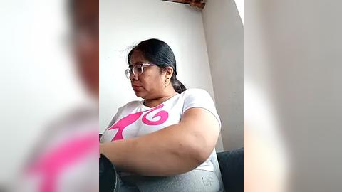 Media: Video of a plus-size Latina woman with medium skin tone, long black hair, and glasses, wearing a gray T-shirt with a pink cat design and gray pants, sitting on a dark blue couch in a simple, bright room.