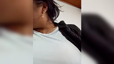 Media: A video showing a woman with medium brown skin and dark hair, wearing a white top and gold earrings, adjusting her hair, with a blurred background.