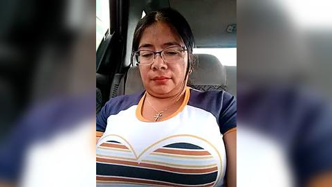Media: Video of a middle-aged woman with glasses and dark hair, wearing a white shirt with horizontal stripes, sitting in a car. Her expression is neutral.