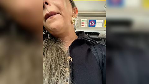 Media: A video shows a middle-aged woman with light skin and gray hair, wearing a black jacket and gold earrings, sitting in a public bus. The background features a blurred bus interior with a yellow and blue sign.