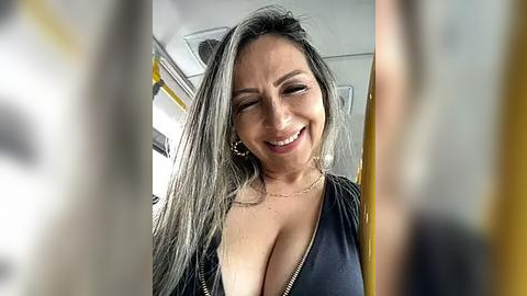 Media: Video of a smiling, middle-aged woman with long, gray hair, wearing a low-cut black top, showing cleavage. She is in a public bus, with yellow seats and windows visible in the blurred background.