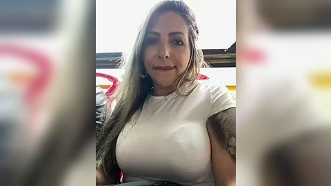 Media: Video of a Latina woman with long, wavy brown hair, wearing a tight white top, revealing her large breasts and tattooed arms, sitting in a car with blurred, colorful background.