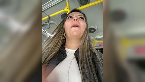 Media: Video of a woman with long, straight, light brown hair, wearing a black jacket over a white top, standing on a yellow-handrail subway car.