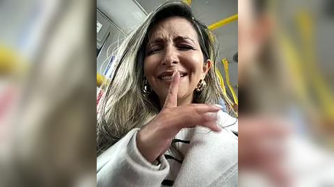 Media: Video of a middle-aged woman with long, straight hair, wearing a white sweater, and large gold hoop earrings, smiling and covering her mouth with her fingers in a public bus.