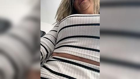 Media: Video of a woman with blonde hair, wearing a black and white striped ribbed crop top, smiling slightly. The background is blurred, with vertical stripes.