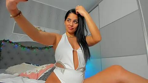 Media: Video of a Latina woman with long black hair, wearing a revealing white one-piece swimsuit, posing playfully on a bed in a modern bedroom with grey walls and festive string lights.