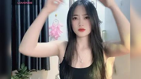 Media: Video of an East Asian woman with long black hair, wearing a black sleeveless top, adjusting her hair, in a room with dark curtains and a dreamcatcher wall hanging.