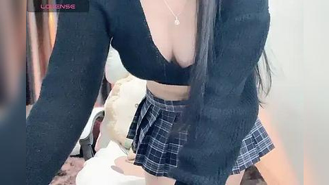 Media: Video of a woman with long black hair, wearing a low-cut black crop top and a short plaid skirt, sitting on a bed, indoors.