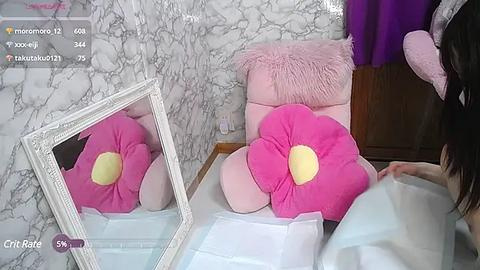 Media: Video of a young woman with long black hair, lying on a pink fluffy chair in a marble-patterned room, surrounded by pink flower pillows and a mirror.