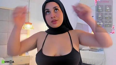 Media: Video of a light-skinned woman with a curvaceous figure and large breasts, wearing a black hijab and revealing tank top, captured mid-dance in a brightly lit bedroom.