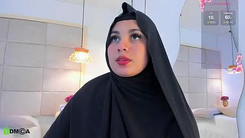 Media: Video of a woman with a medium skin tone wearing a black hijab and red lipstick, in a modern, bright bathroom with tiled walls, hanging lights, and a bathtub.