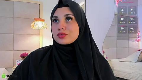 Media: Video of a Middle Eastern woman with fair skin, wearing a black hijab, standing in a modern bedroom with beige walls, white bed, and a calendar.