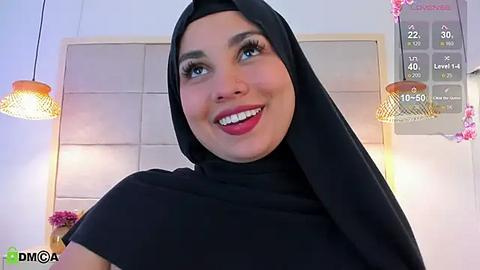 Media: Video of a smiling woman with fair skin, wearing a black hijab and red lipstick, in a modern room with a beige padded headboard and hanging lamps.