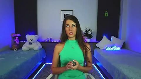 Media: Video of a young woman with long brown hair, wearing a green sleeveless top, standing in a dimly-lit bedroom with two twin beds, teddy bears, and neon lights.