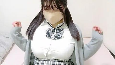 Media: Video of an Asian woman with long black hair in pigtails, wearing a white blouse, gray cardigan, and plaid skirt, posing with fists raised in a playful manner against a plain, light-colored background.