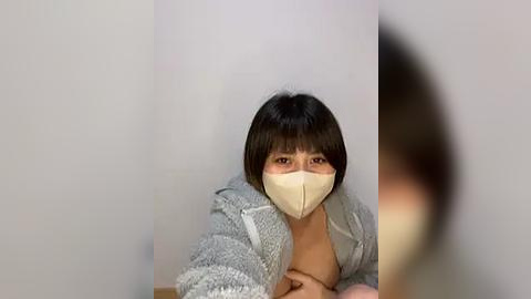 Media: A video of a woman with short black hair, wearing a beige surgical mask and a light gray, fuzzy cardigan, standing against a plain, light-colored wall. The image is slightly blurred, focusing on her face.