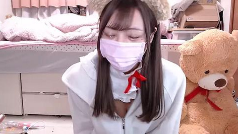 Media: Video of a young Asian woman with long black hair, wearing a light blue hoodie and a white surgical mask, kneeling in a cluttered, cozy bedroom with a teddy bear and a bed with pink sheets.