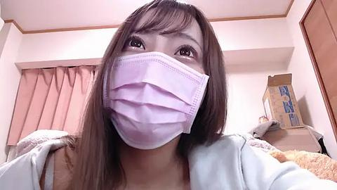 Media: A video of an East Asian woman with long brown hair, wearing a light blue surgical mask, in a cluttered bedroom with beige walls and pink curtains.