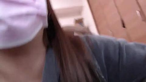 Media: A video of a person with long, brown hair wearing a white face mask, a gray sweater, and standing in front of a wooden door. The image is slightly blurry.