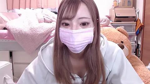 Media: Video of a young East Asian woman with light skin and long, straight brown hair, wearing a white surgical mask, sitting in a cluttered bedroom with a teddy bear and various personal items.