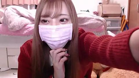 Media: Video of a young Asian woman with straight brown hair, wearing a red sweater, white mask, and black hairband, kneeling in a messy bedroom with pink blankets and a stuffed bear.