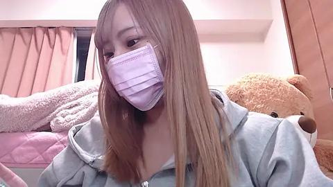 Media: Video of an East Asian woman with long, straight, light brown hair, wearing a pink surgical mask, light gray hoodie, and a plush teddy bear in a cozy, pastel-toned bedroom.