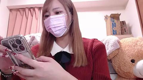 Media: Video of a young Asian woman with long straight hair wearing a pink face mask, red sweater, and black bow tie, holding a smartphone in a cozy room with beige curtains, stuffed animals, and cardboard boxes in the background.