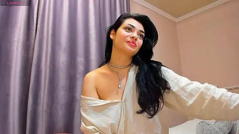 Media: Video of a smiling woman with long black hair, wearing a white off-shoulder shirt and multiple necklaces, standing in a softly lit bedroom with lavender curtains and beige walls.