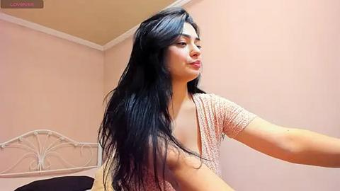 Media: Video of a young woman with long, black hair and light skin, wearing a pink patterned top, standing in a peach-colored room with a white metal bed frame.