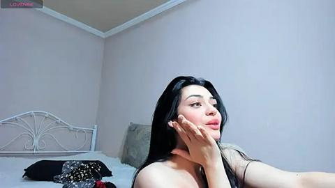 Media: Video of a young Asian woman with long black hair, light skin, and a slender build, resting her chin on her hand in a bedroom with white walls and a metal bed frame.