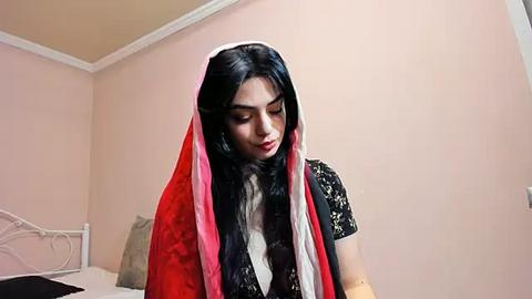 Media: A video of a young woman with long black hair, wearing a traditional red and black sari, standing in a simple beige-walled bedroom with a bed in the background.