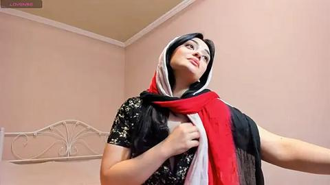 Media: Video of a young woman with fair skin and black hair, wearing a black floral dress and a red and white scarf, standing in a beige-walled room with a white metal headboard.