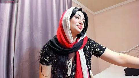 Media: Video of a light-skinned woman with long black hair, wearing a black floral top, a red and white headscarf, and a red scarf, smiling, in a beige room with a purple curtain.