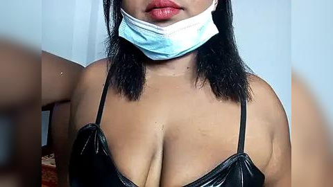 Media: Video of a woman with medium brown skin, wearing a black latex bra and a blue surgical mask, with straight black hair.