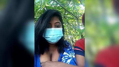 Media: Video of a Black woman with medium-dark skin, wearing a blue face mask, blue top, and black hair, outdoors among lush green foliage.