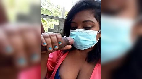 Media: A video of a South Asian woman with dark skin, wearing a blue surgical mask, blue nail polish, and a red jacket, holding a large erect penis with her right hand.