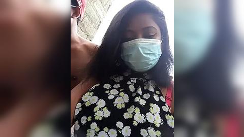 Media: Video of a woman with long black hair, wearing a black floral dress, light blue surgical mask, and red sunglasses. Background shows a blurry brick wall.