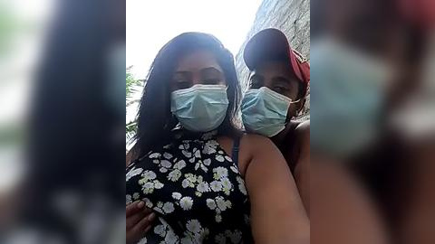 Media: Video of two Black women wearing masks, one with a floral top, the other in a cap, standing in front of a brick wall, with blurred greenery in the background.