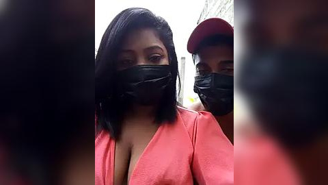 Media: Video of two Black women with medium brown skin, wearing black masks, pink blouses, and a red cap, standing outdoors.