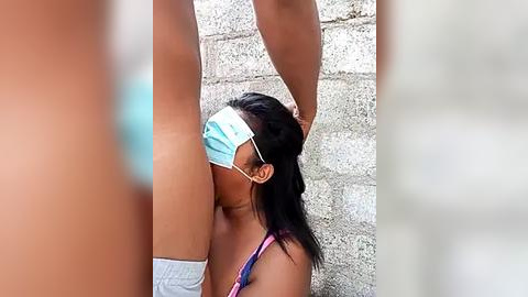 Media: Video of a dark-skinned woman with a mask, kneeling before a man's raised arm. She wears a bra, revealing her breasts, against a concrete wall. The image is slightly blurred.