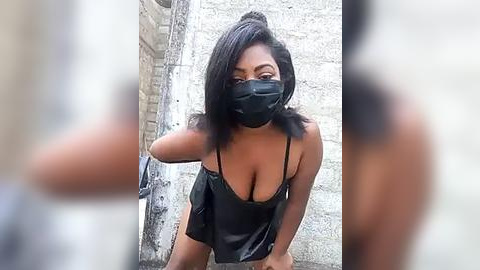 Media: Video of a Black woman with medium-dark skin tone, wearing a black face mask, black spaghetti-strap tank top, and black panties, posing provocatively against a rough concrete wall.