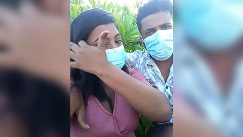 Media: A video captures a young Black woman with long, straight black hair, wearing a pink top, and a man with short curly hair, both wearing blue surgical masks, standing closely together in a lush green outdoor setting.