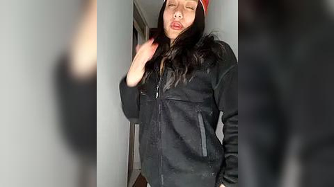 Media: Video of an Asian woman with long black hair and a red beanie, wearing a black, zip-up jacket, standing in a dimly lit hallway. She appears to be making a kissing gesture with her lips.