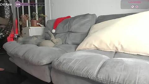 Media: Video of a plush gray sectional sofa with a red Santa hat and beige pillow, against a white wall with a window and greenery.