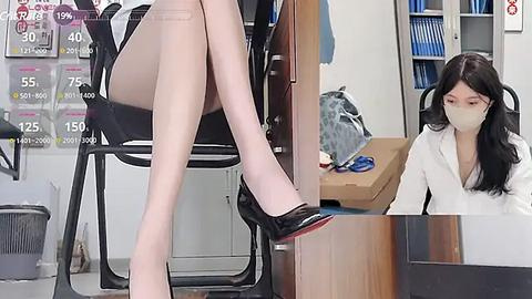 Media: Video of an office scene: a woman in a white blouse sits at a desk, wearing a mask, while a legged mannequin with sheer tights and black heels stands beside her.