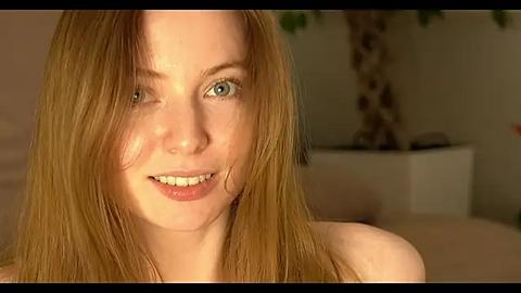 Media: Video of a young, smiling woman with fair skin and long, straight, light brown hair. She has blue eyes and is wearing a sleeveless top. Background includes a blurred indoor setting with a potted plant.