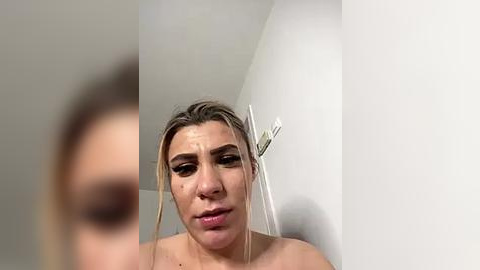 Media: Video of a young woman with blonde hair and light skin, looking distressed, standing in a white bathroom with a showerhead.